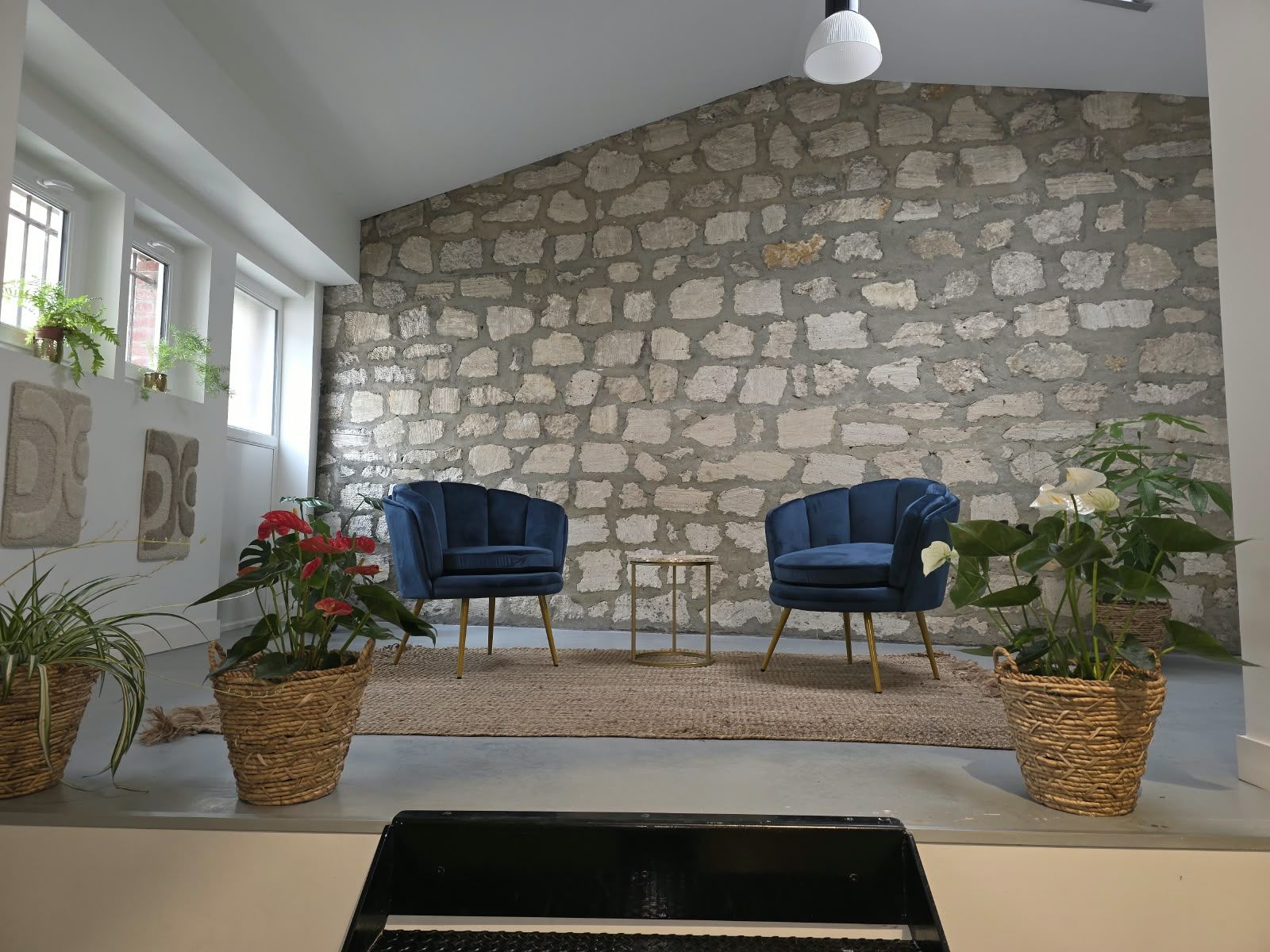 L’estrade : deux fauteuils en velours bleu nuit et une table d’appoint dorée devant un mur en pierre, entourés de plantes